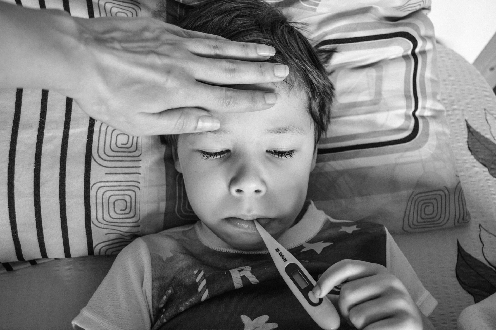 Dijete leži u krevetu s toplomjerom, roditelj mu drži ruku na čelu